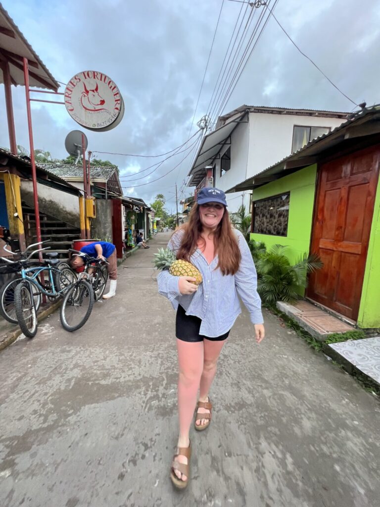 Smiling girl holding pineapple standing in alley of colorful buildings.