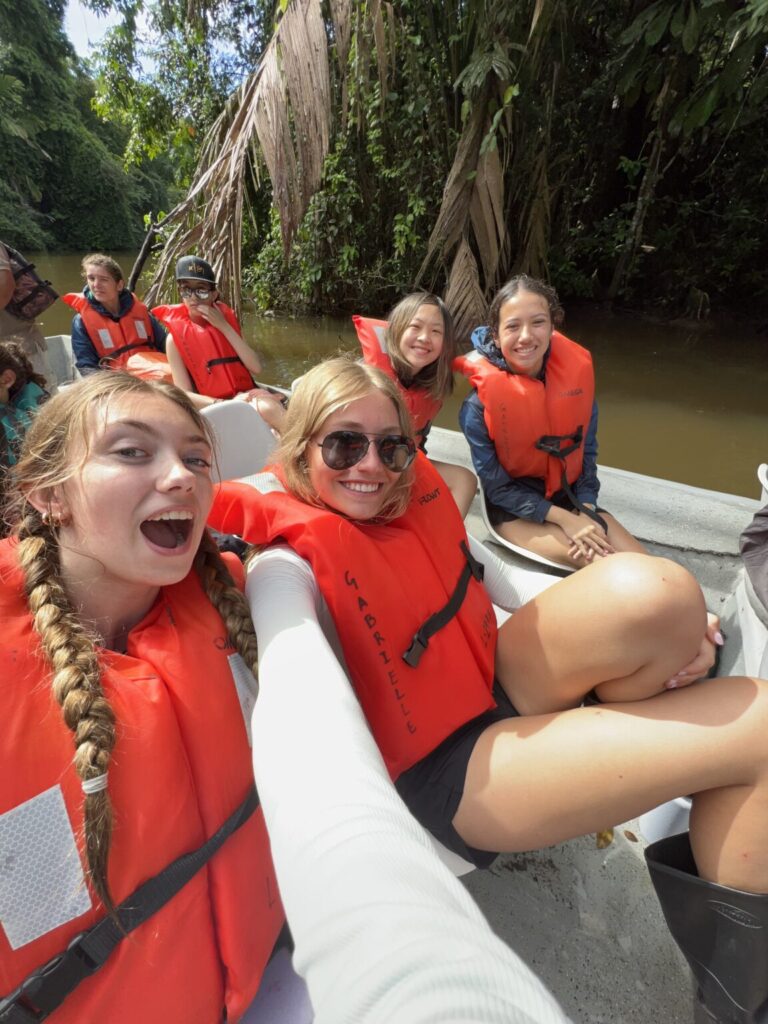 Students taking selfie while wearing orange life vests and sitting on boat.