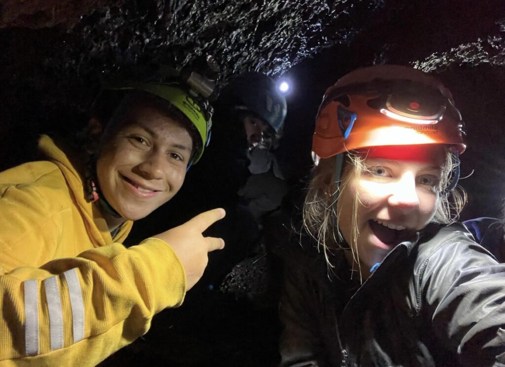 Two students wearing hard hats taking selfie in dark cave.