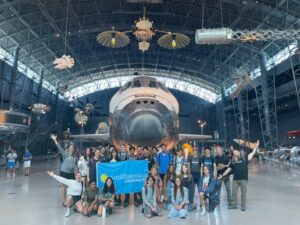 Group of students holding blue Smithsonian Journeys flag in front of a large plane.