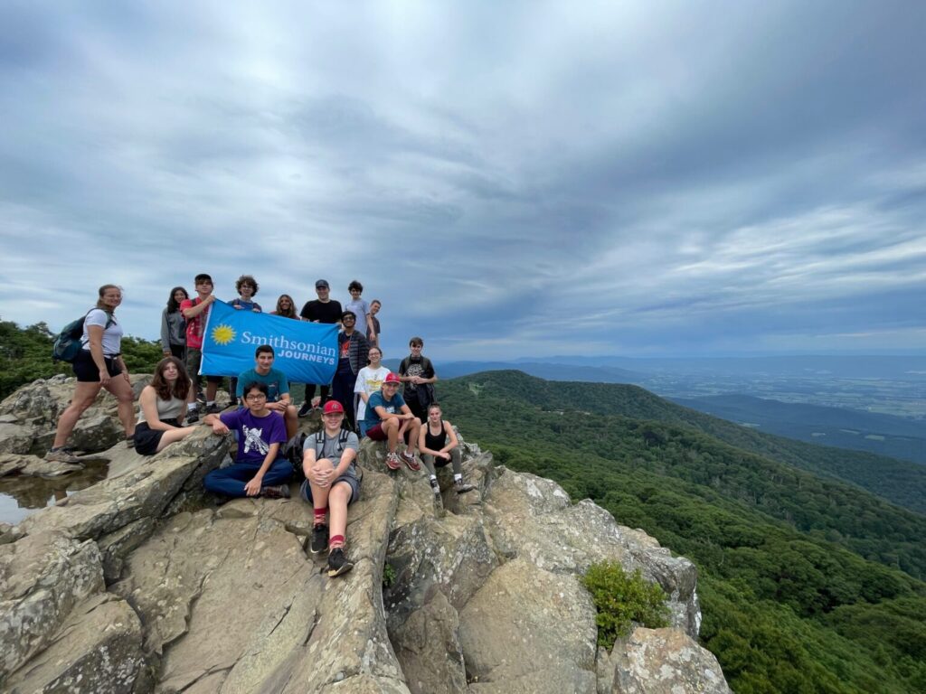 Students holding blue Smithsonian Journeys flag on top of mountain.