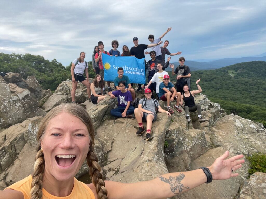 Woman smiling in front of group of students sitting on top of mountain.