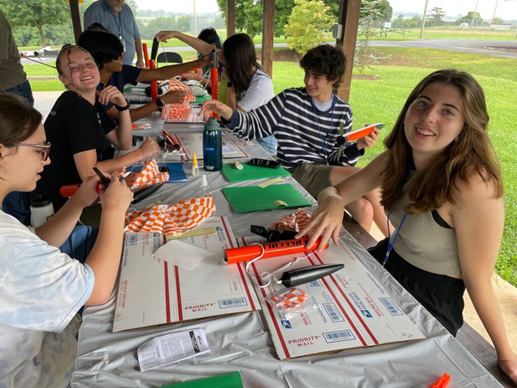 Students sitting at table outside with rocket parts. 